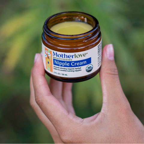 Hand holding a jar of Motherlove Nipple Cream against a blurred green background
