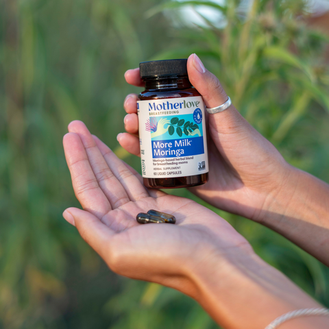 Hand holding a bottle of Motherlove More Milk Moringa supplement with a blurred green background
