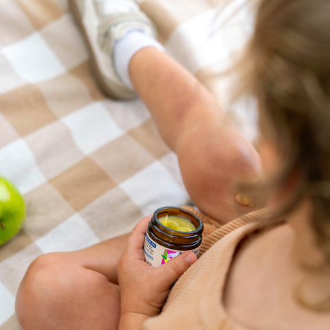 Child holding a jar of Baby Hand and Knee Balm with a green apple on a checkered blanket