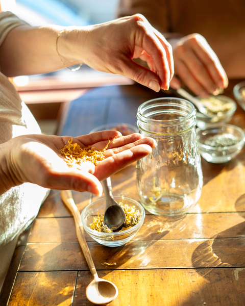Person holding a small amount of dried calendula flowers over a glass jar on a wooden table.