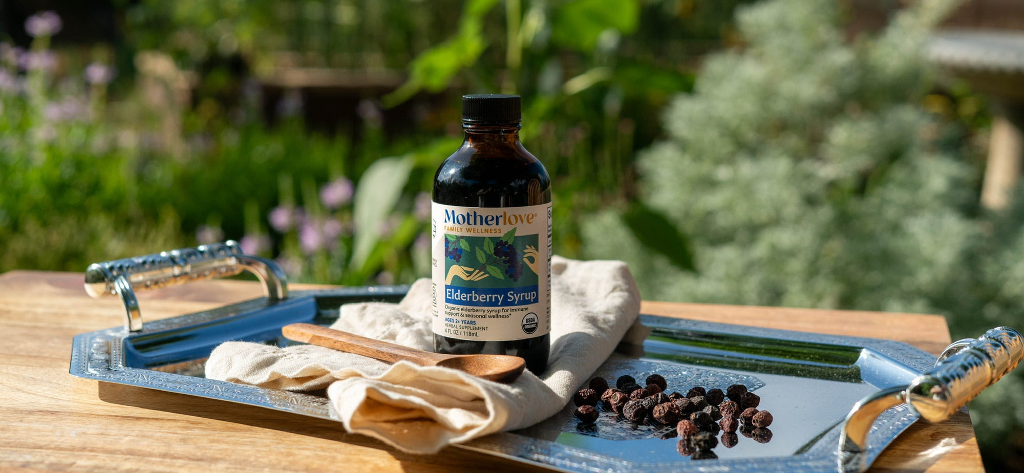 A bottle of Motherlove Blueberry Syrup sits on a rustic metal tray on a wooden table, with berries, a cloth, and a wooden spoon nearby, set against a blurred outdoor garden background.