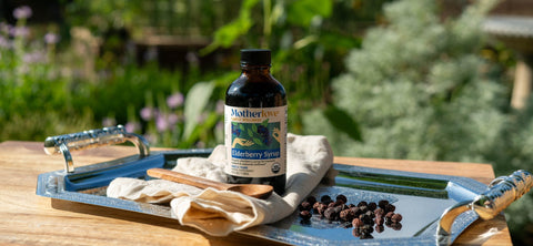 A bottle of Motherlove Blueberry Syrup sits on a rustic metal tray on a wooden table, with berries, a cloth, and a wooden spoon nearby, set against a blurred outdoor garden background.
