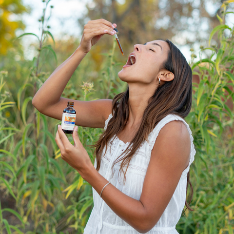Woman in a white dress outdoors, holding a bottle and dropper near her mouth.