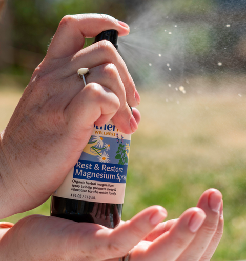 Person holding a bottle of 'Rest & Restore Magnesium Spray' and spraying it onto their hands outdoors.