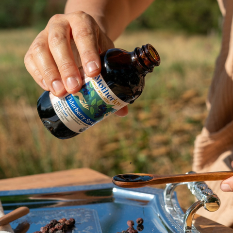 A hand holds an amber glass bottle labeled Mother of Elderberry Syrup over a metal tray with a wooden spoon, outdoors with blurred grass in the background.