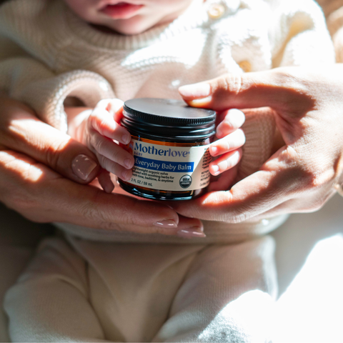 Person holding a jar of Motherlove Everyday Baby Balm with a baby in the background.
