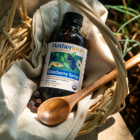 A bottle of Mother Love Family Wellness Elderberry Syrup placed in a woven basket with a wooden spoon and cloth, outdoors on a stone surface with natural light and shadows.