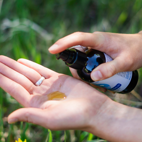 A person outdoors in a grassy area pumps a dark bottle onto their open palm, showing a small amount of amber-colored liquid. The person is wearing a silver ring on their left hand.