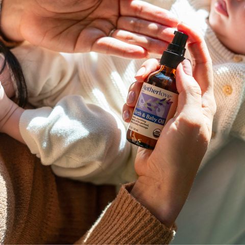 Person holding a bottle of Motherlove Birth & Baby Oil with a child in the background