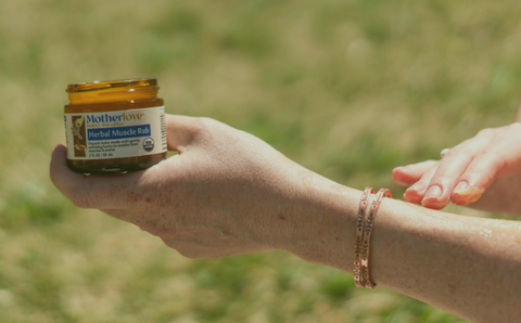 Person holding a jar of Motherlove Herbal Muscle Balm on a blurred green background