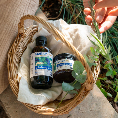 A wicker basket lined with cream fabric holds two Mother brand products, a dark amber bottle labeled Elderberry Soap and a dark jar labeled Children's chest rub alongside green leaves. A person's hand and torso are visible in a natural outdoor setting.